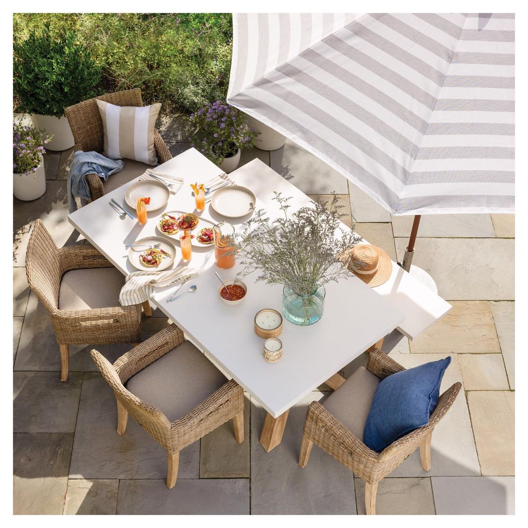 Outdoor dining setup features a square white table with wicker chairs, place settings, drinks, and a striped patio umbrella for shade.