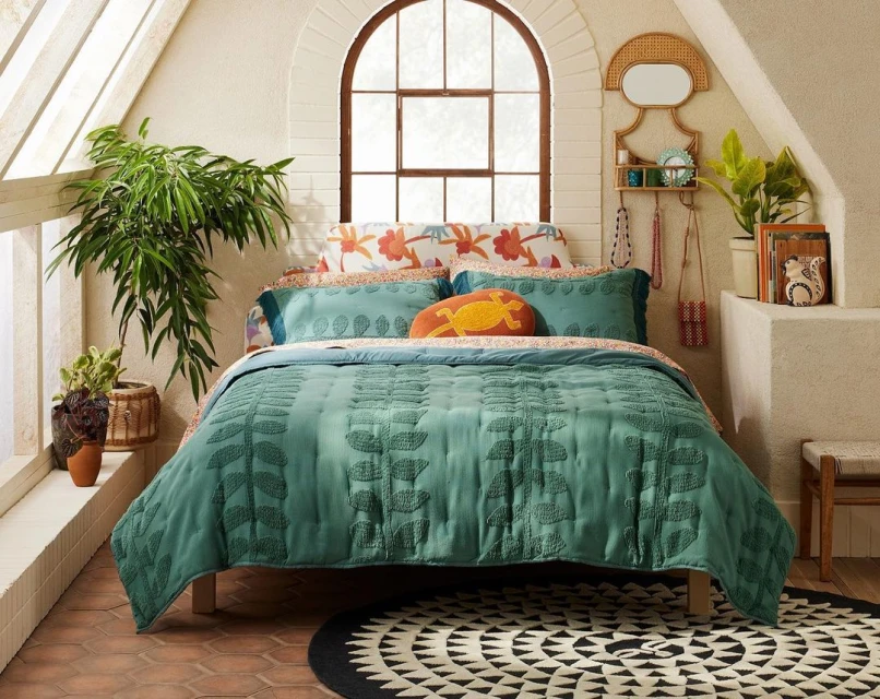 Boho bedroom with teal bedspread featuring embroidered leaf pattern, bright floral pillows, houseplants, round rug and arched window.