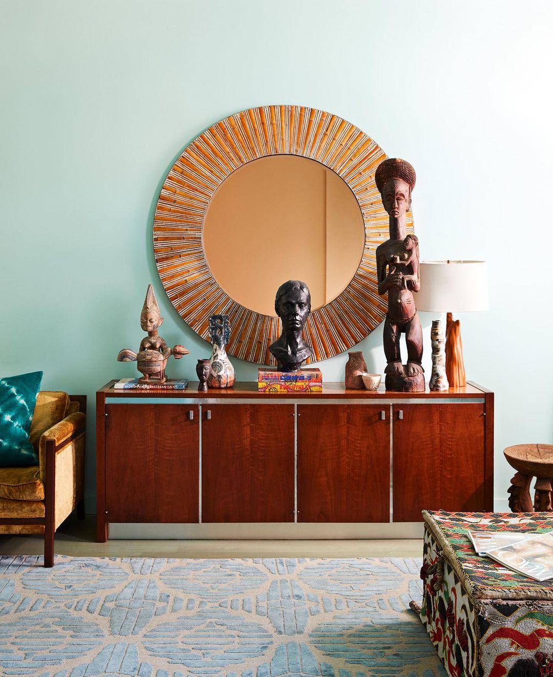 Interior design featuring a wooden sideboard with decorative objects, a round mirror, and patterned rug against a light blue wall.