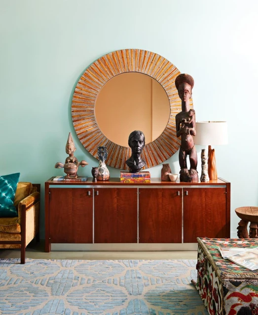 Interior design featuring a wooden sideboard with decorative objects, a round mirror, and patterned rug against a light blue wall.