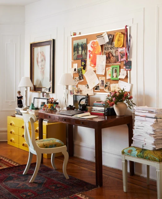 Home office interior with a wooden desk cluttered with books and decor, a white chair, and a corkboard on a white wall.