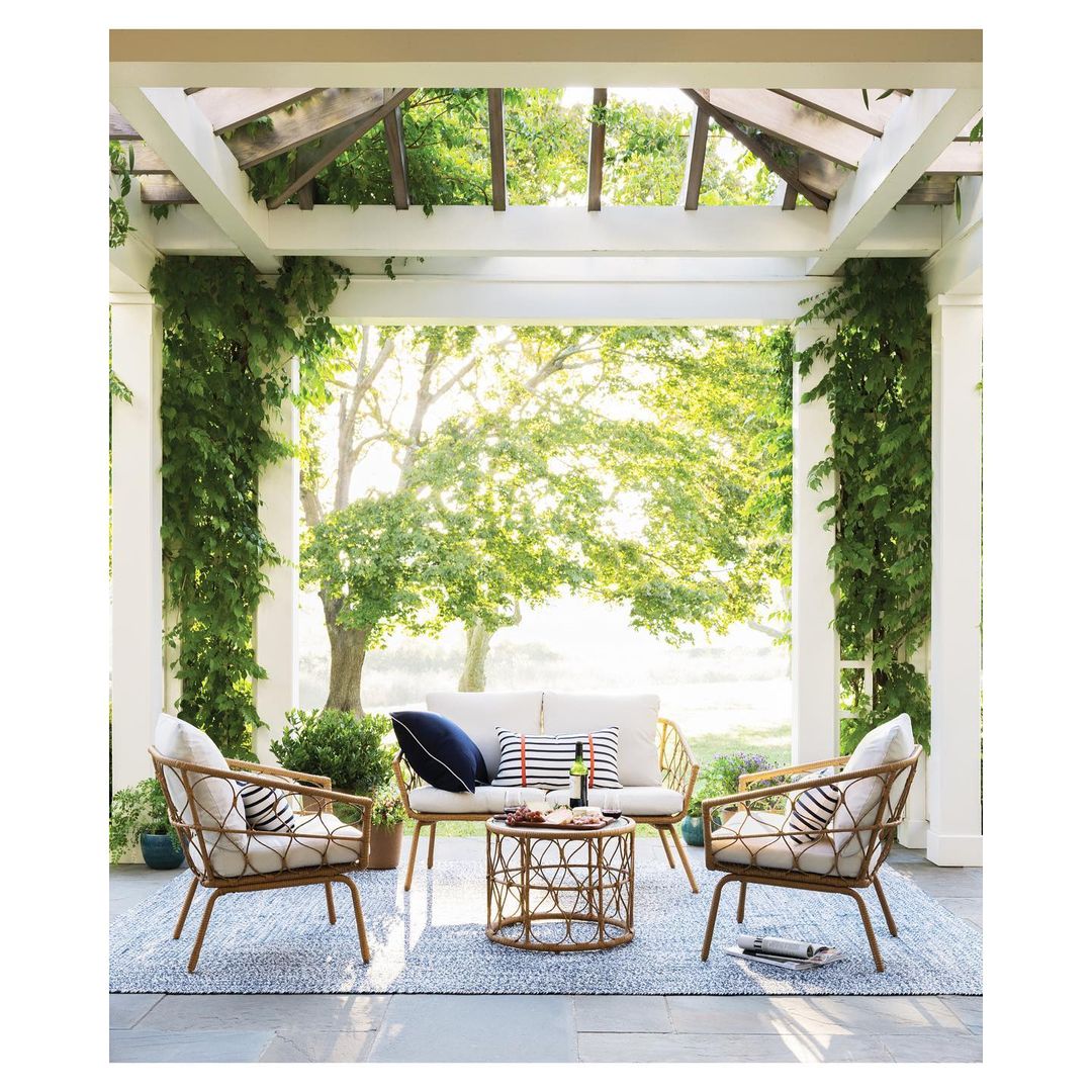 Outdoor patio setup with rattan furniture under a pergola, featuring a sofa, chairs, and a coffee table on a patterned blue rug.