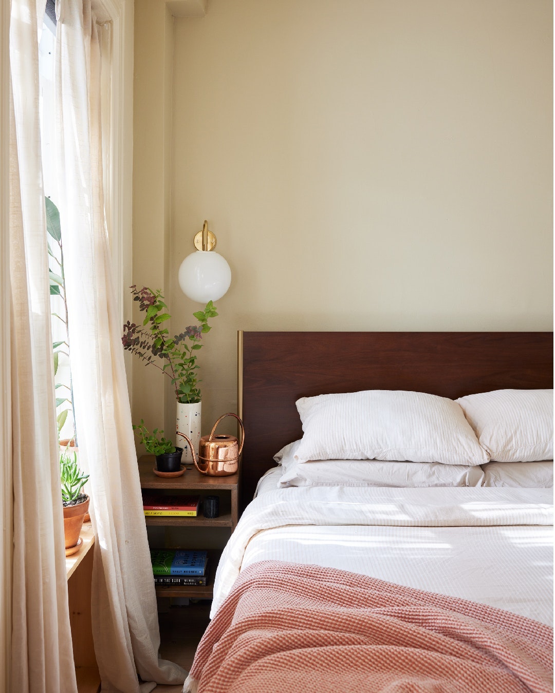 Bedroom with a queen-sized bed, nightstand, sheer curtains, and indoor plants in a warm, inviting interior space with soft, natural light.