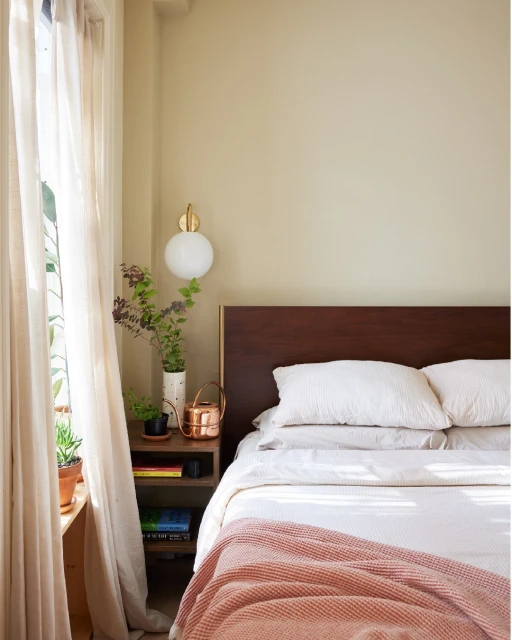 Bedroom with a queen-sized bed, nightstand, sheer curtains, and indoor plants in a warm, inviting interior space with soft, natural light.
