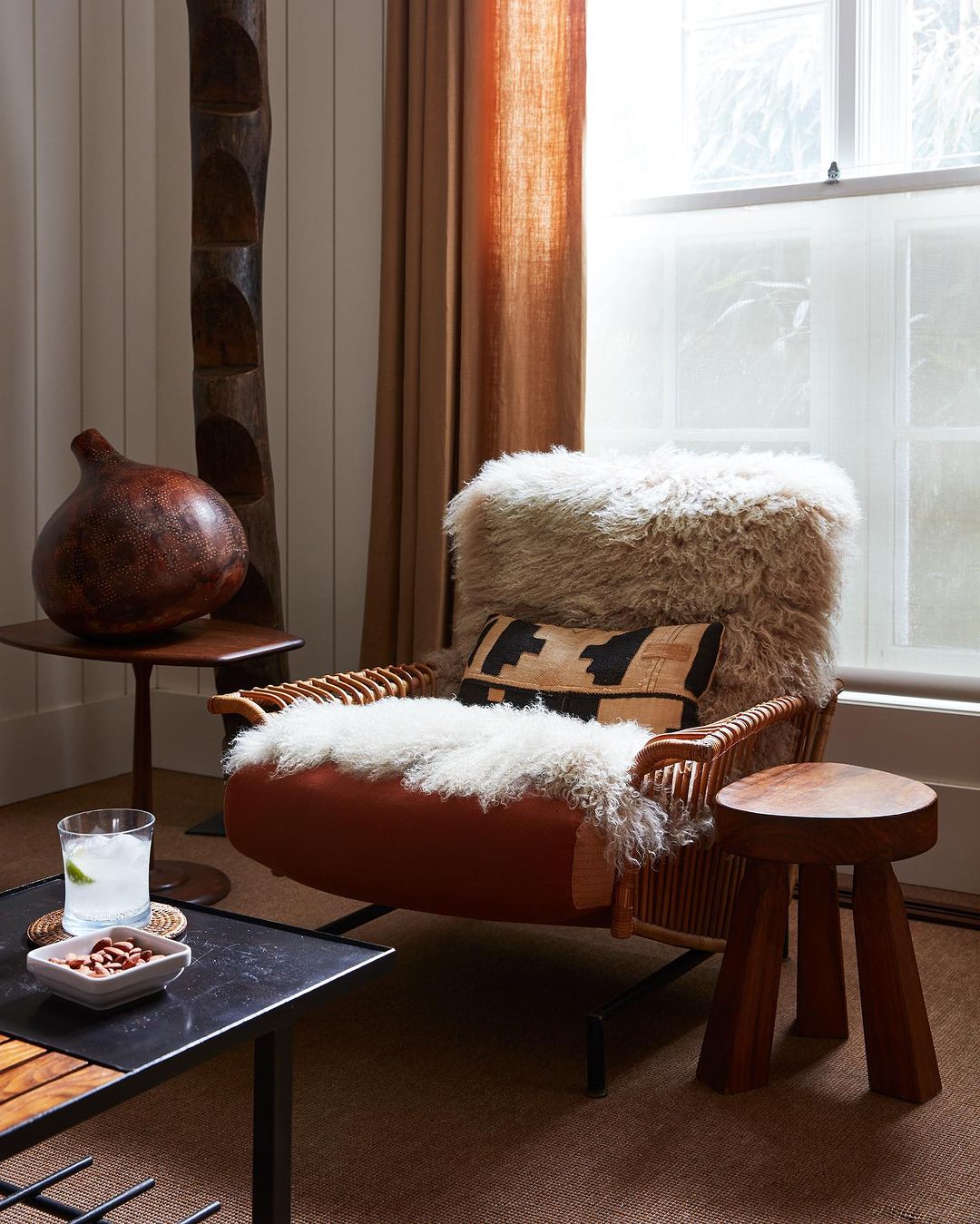 Living room corner featuring a wicker armchair with a sheepskin throw, a geometric cushion, and a dark wood side table.