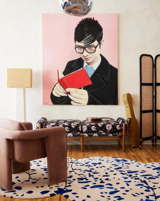 Painting of a person wearing glasses and a black coat, reading a red book, against a pink background, in a modern interior setting.
