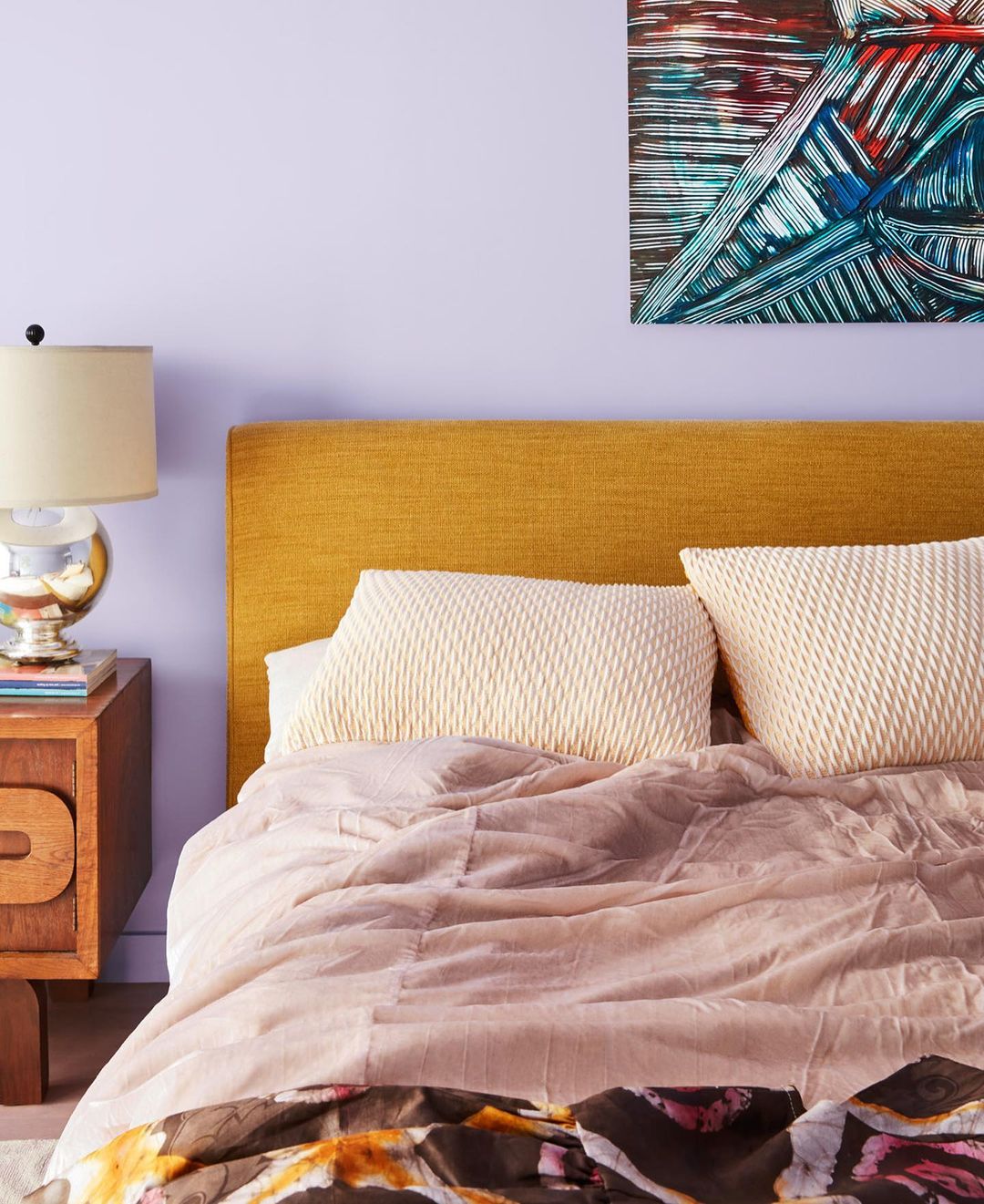 Bedroom interior featuring a bed with patterned bedding, a mustard-colored headboard, nightstand with lamp, and abstract artwork.