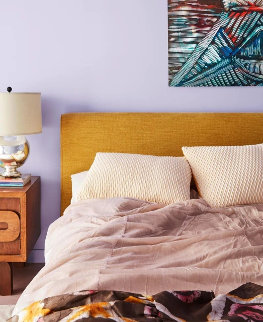 Bedroom interior featuring a bed with patterned bedding, a mustard-colored headboard, nightstand with lamp, and abstract artwork.