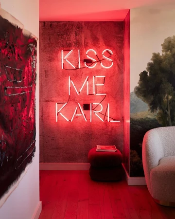 Interior with an illuminated 'Kiss Me Karl' neon sign on a textured wall, featuring modern decor, an abstract painting, and warm lighting.