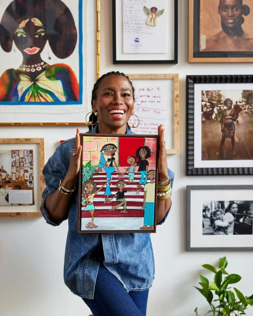 Smiling African-American woman holds a colorful painting of a family; wall with framed art creates an indoor, home studio backdrop.