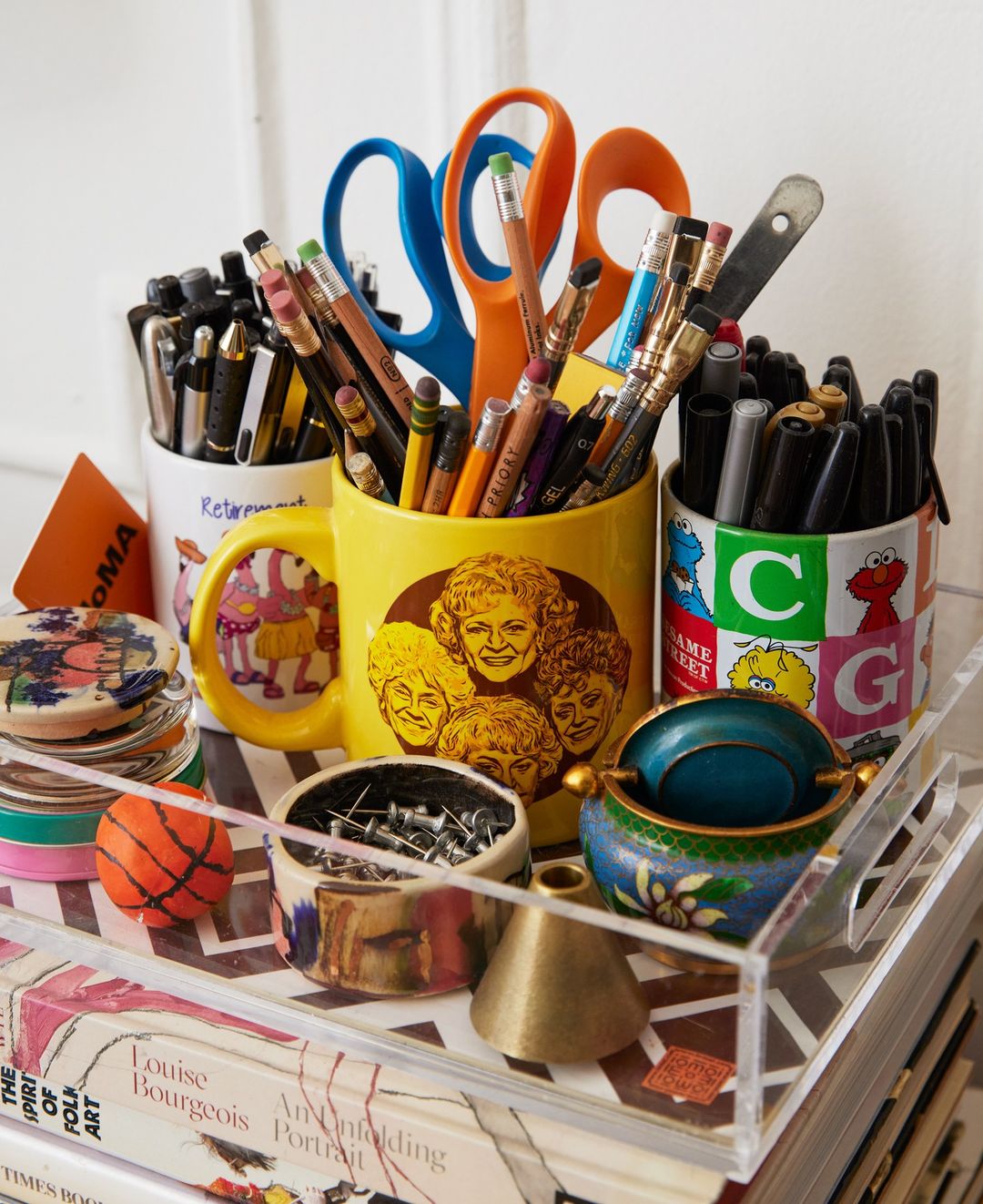 Collection of pens, pencils, scissors, pins, and small objects held in decorated mugs and small containers, stacked on books.
