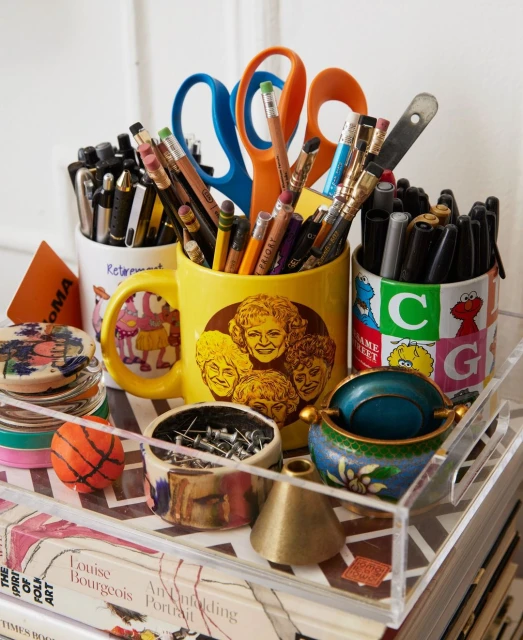 Collection of pens, pencils, scissors, pins, and small objects held in decorated mugs and small containers, stacked on books.