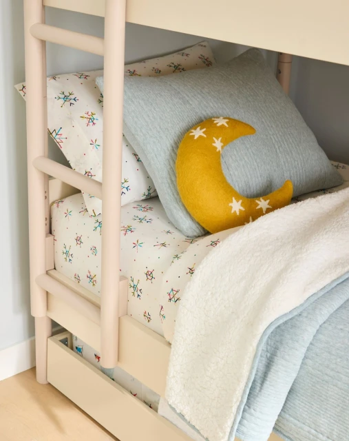 Bunk bed with patterned sheets, soft gray pillow, moon-shaped pillow with stars, and soft white blanket in a brightly lit interior.