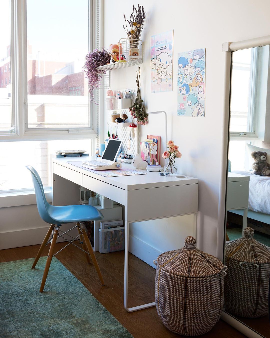 Bright, minimalist home office setup featuring a white desk, blue chair, laptop, flowers, and decorative storage with natural light from a large window.