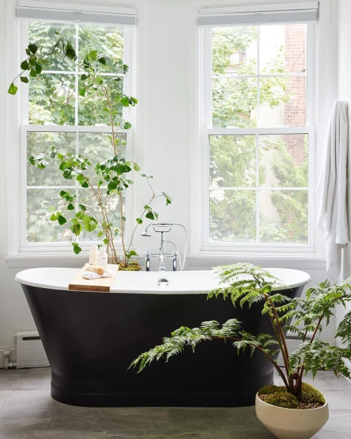 Contemporary bathroom with a black freestanding bathtub, white walls, two windows, potted plants, and natural light.