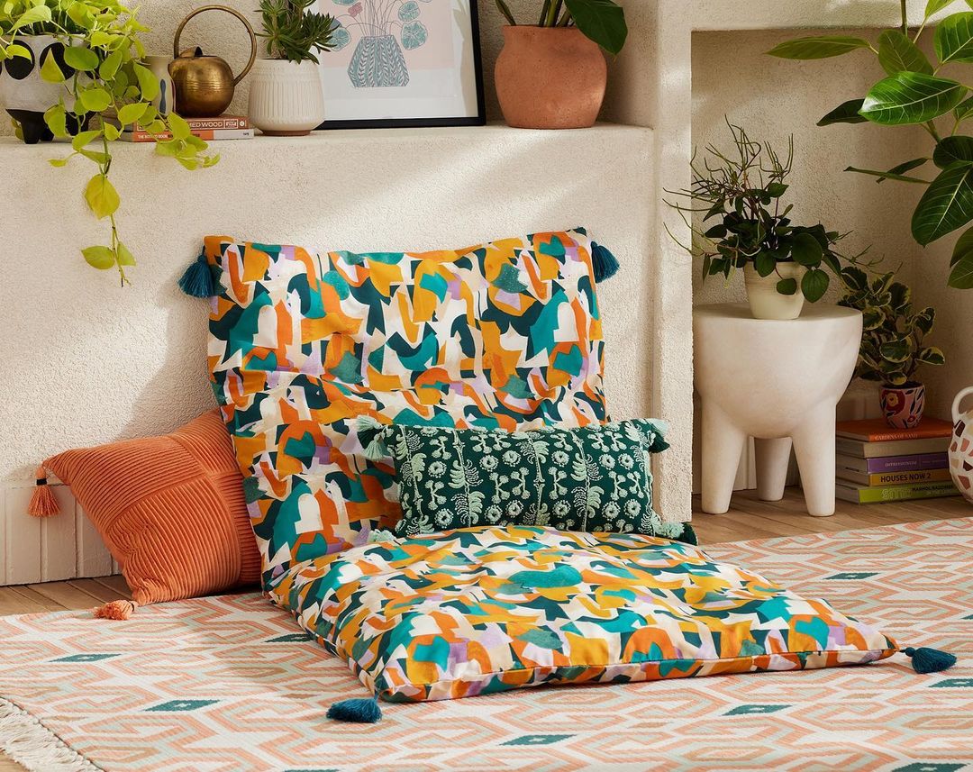 Interior living room with floor cushions and throw pillows on patterned rug, with houseplants and decor on shelves.