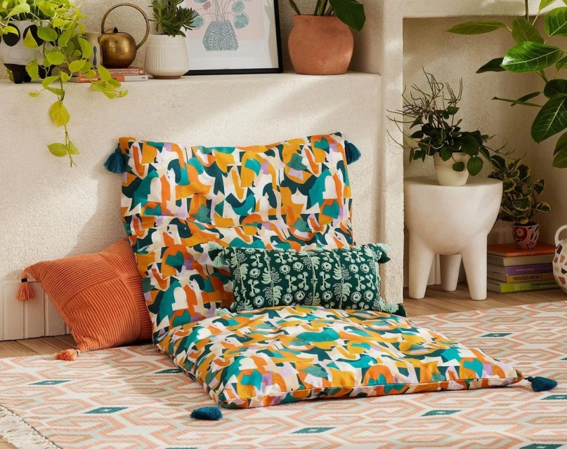 Interior living room with floor cushions and throw pillows on patterned rug, with houseplants and decor on shelves. 
