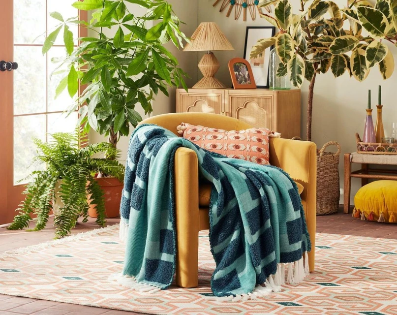 Cozy living room interior featuring a mustard yellow armchair draped with a teal blanket, potted plants, and patterned rug creating a warm space.