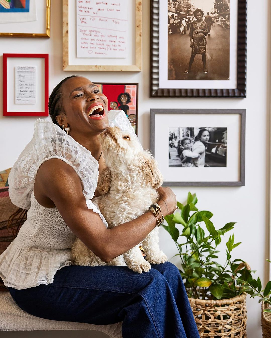 Adult woman laughs while sitting down and holding a dog in her lap with framed pictures on the wall and plants nearby indoors.