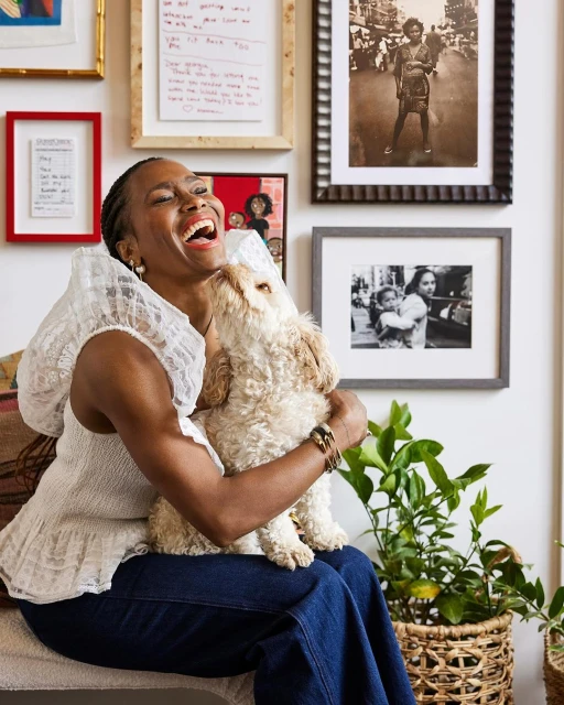 Adult woman laughs while sitting down and holding a dog in her lap with framed pictures on the wall and plants nearby indoors.