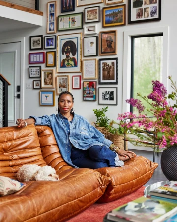 A woman lounges on a leather couch with her dog, in a cozy living room filled with a gallery wall of framed art and photography.