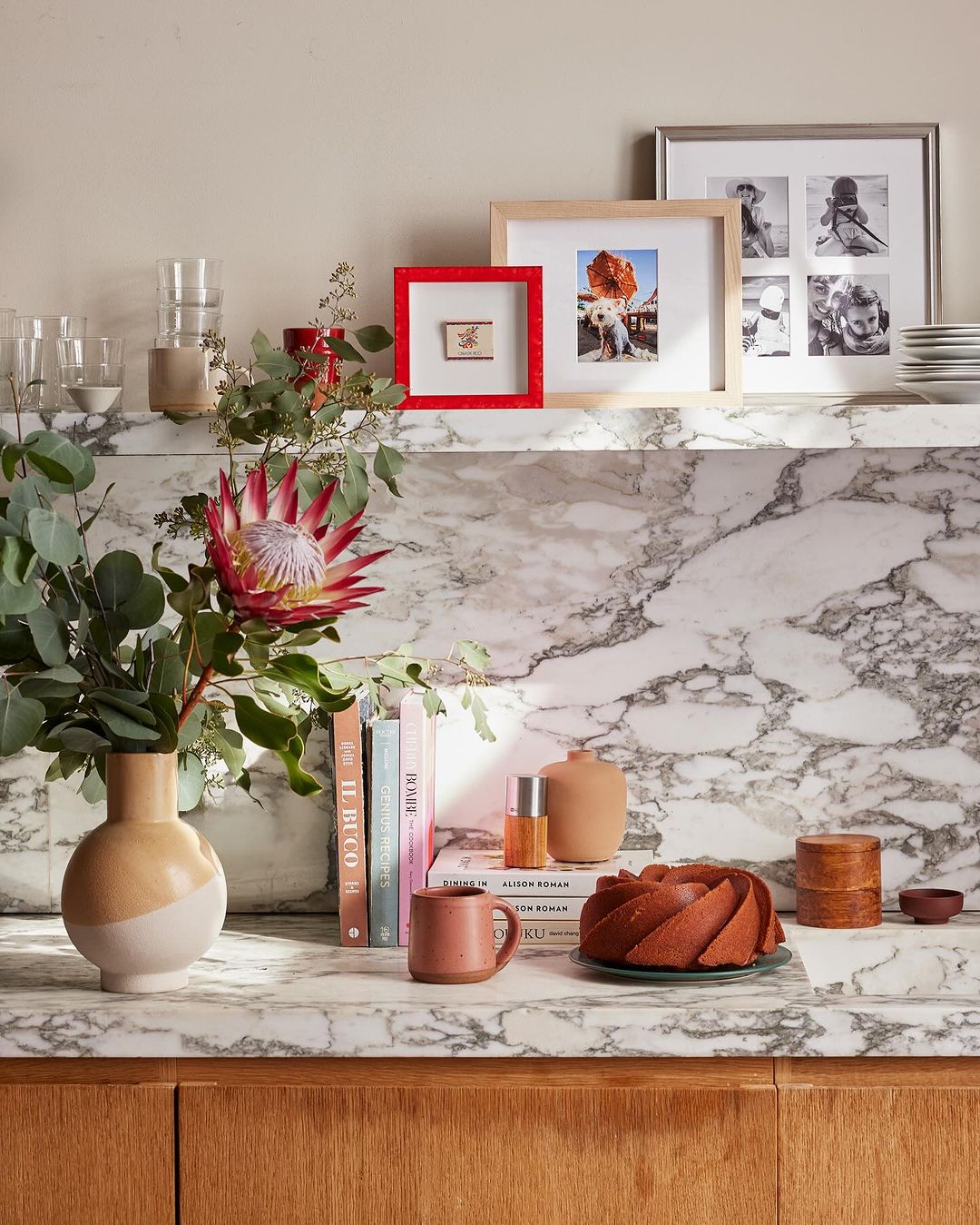 Still life composition featuring a protea flower arrangement, framed photos, and decorative objects on a marble countertop against a neutral wall.