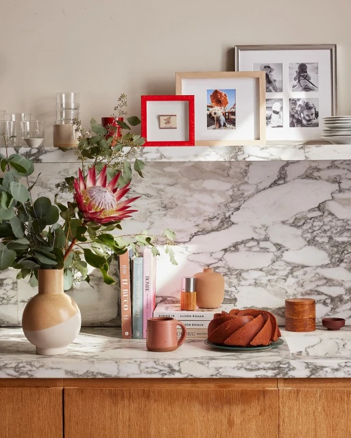 Still life composition featuring a protea flower arrangement, framed photos, and decorative objects on a marble countertop against a neutral wall.