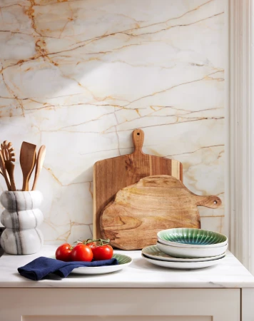 Kitchen scene featuring wooden cutting boards, tomatoes on a plate, ceramic dishes, a marble utensil holder, and a marble backsplash.