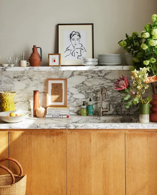 Interior shot of a marble countertop with decor: drawings, vases, flowers, dishes, and books on top of wooden cabinets.