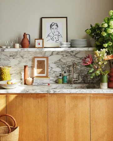 Interior shot of a marble countertop with decor: drawings, vases, flowers, dishes, and books on top of wooden cabinets.