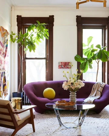 Living room interior with a modern purple couch, round coffee table, potted plants near windows, and decorative wall art.