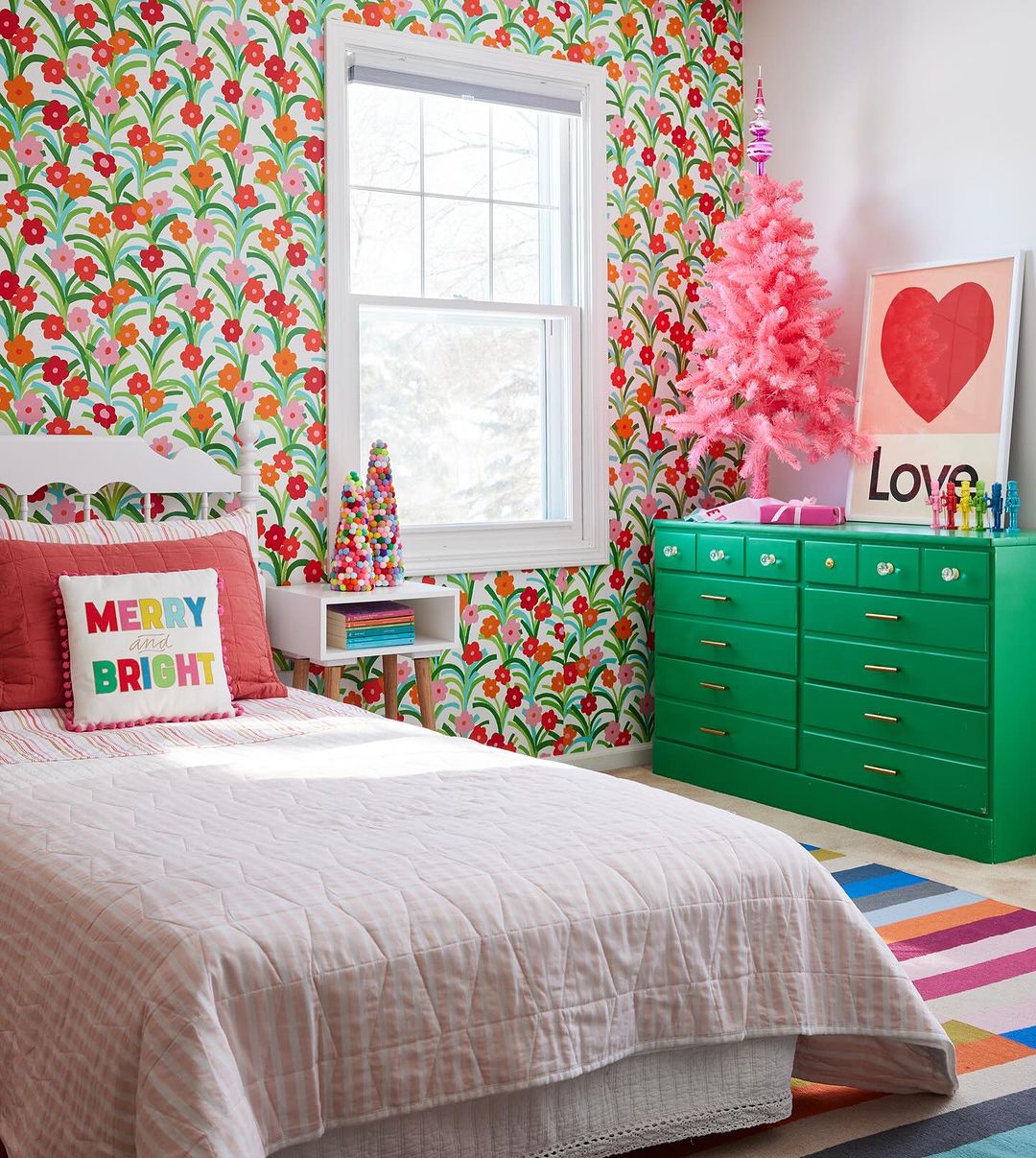 Brightly decorated child's bedroom with bed, green dresser, pink Christmas tree, colorful floral wallpaper, and striped rug.