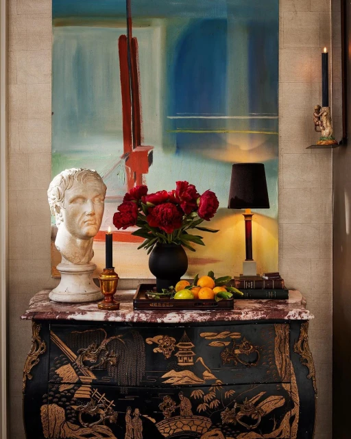 Interior design featuring a black and gold painted chest of drawers, a marble bust, an abstract painting, and red flowers in a vase.
