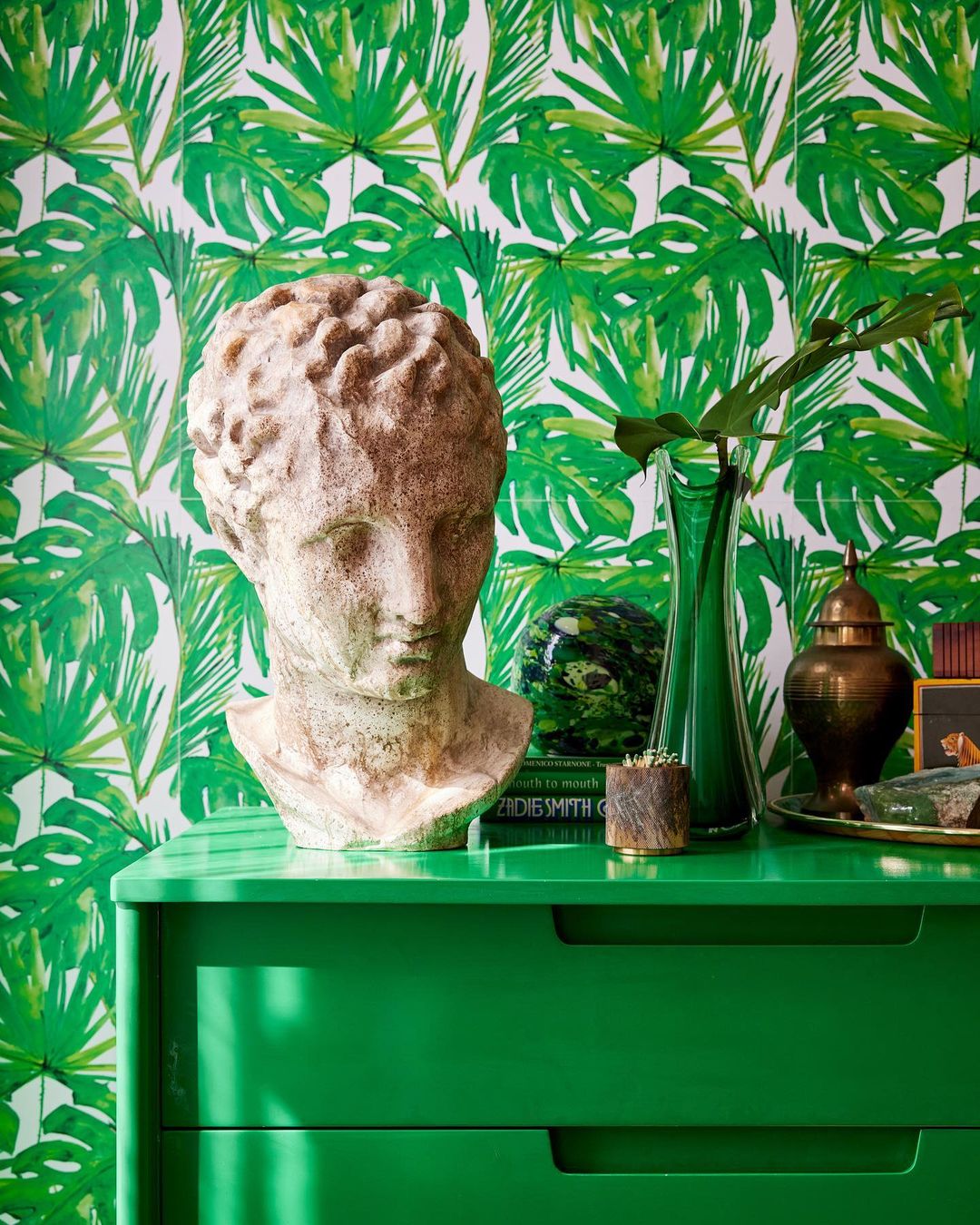 Bust sculpture on a green cabinet against a tropical monstera leaf patterned wall with decorative objects including vases and books.