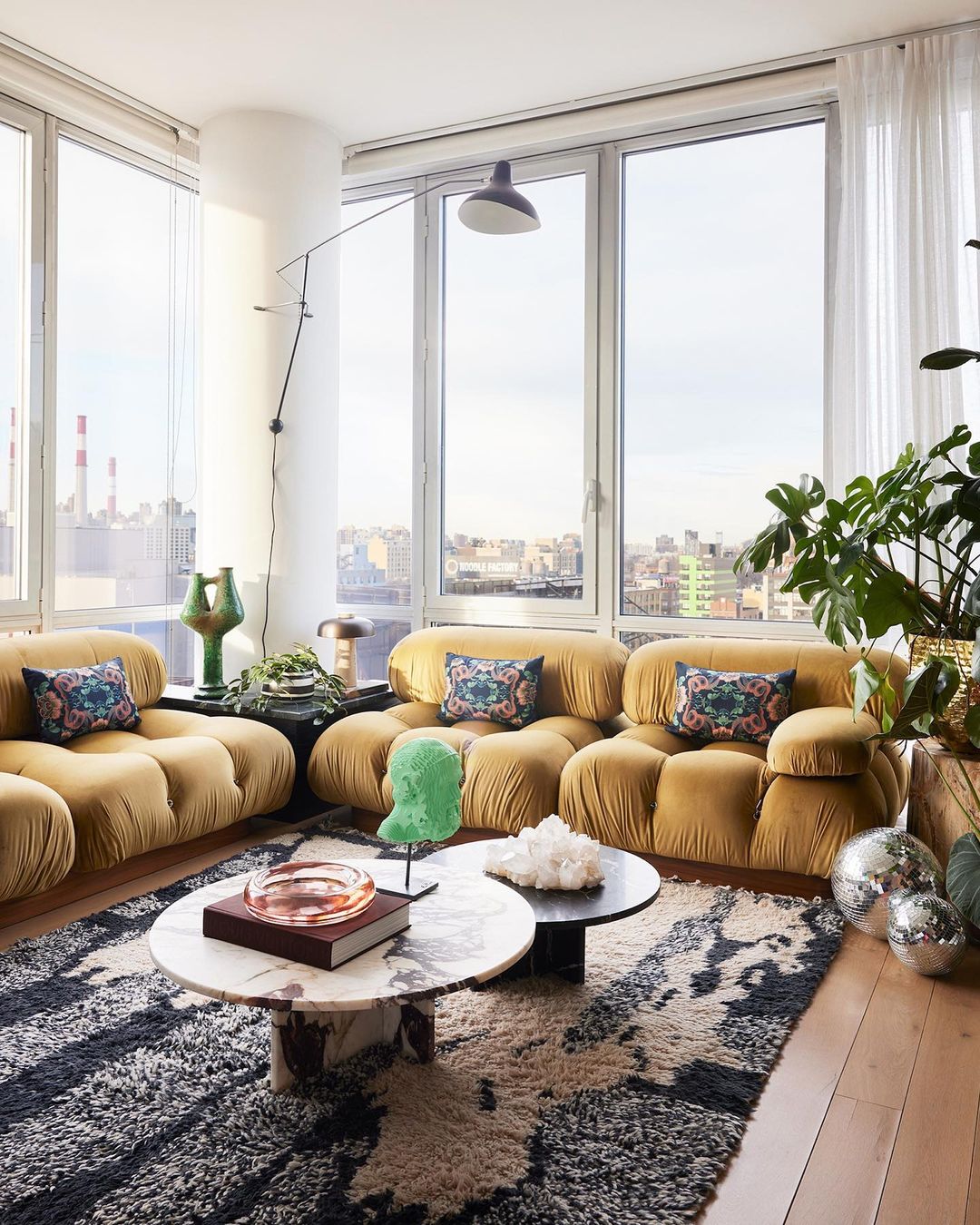 Interior view of a living room featuring a tan tufted sofa, marble coffee tables, a patterned rug, and city views from large windows.