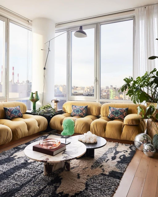 Interior view of a living room featuring a tan tufted sofa, marble coffee tables, a patterned rug, and city views from large windows.