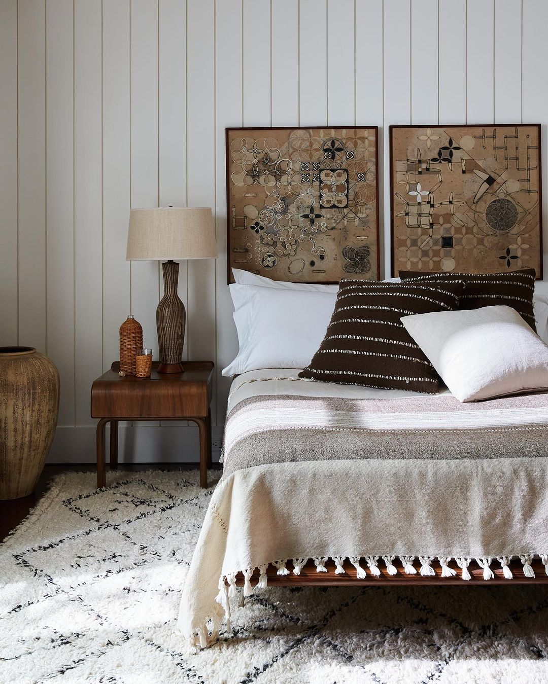 Bedroom interior featuring a bed with neutral-toned bedding, decorative cushions, framed wall art, a wooden side table, and a patterned rug.