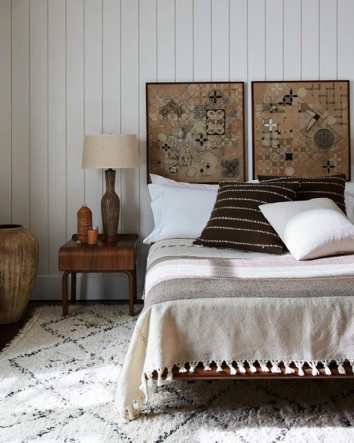 Bedroom interior featuring a bed with neutral-toned bedding, decorative cushions, framed wall art, a wooden side table, and a patterned rug.