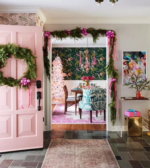 Festive holiday decor in a home entryway featuring a pink door with a wreath, garland, rug, and Christmas decorations in the background.