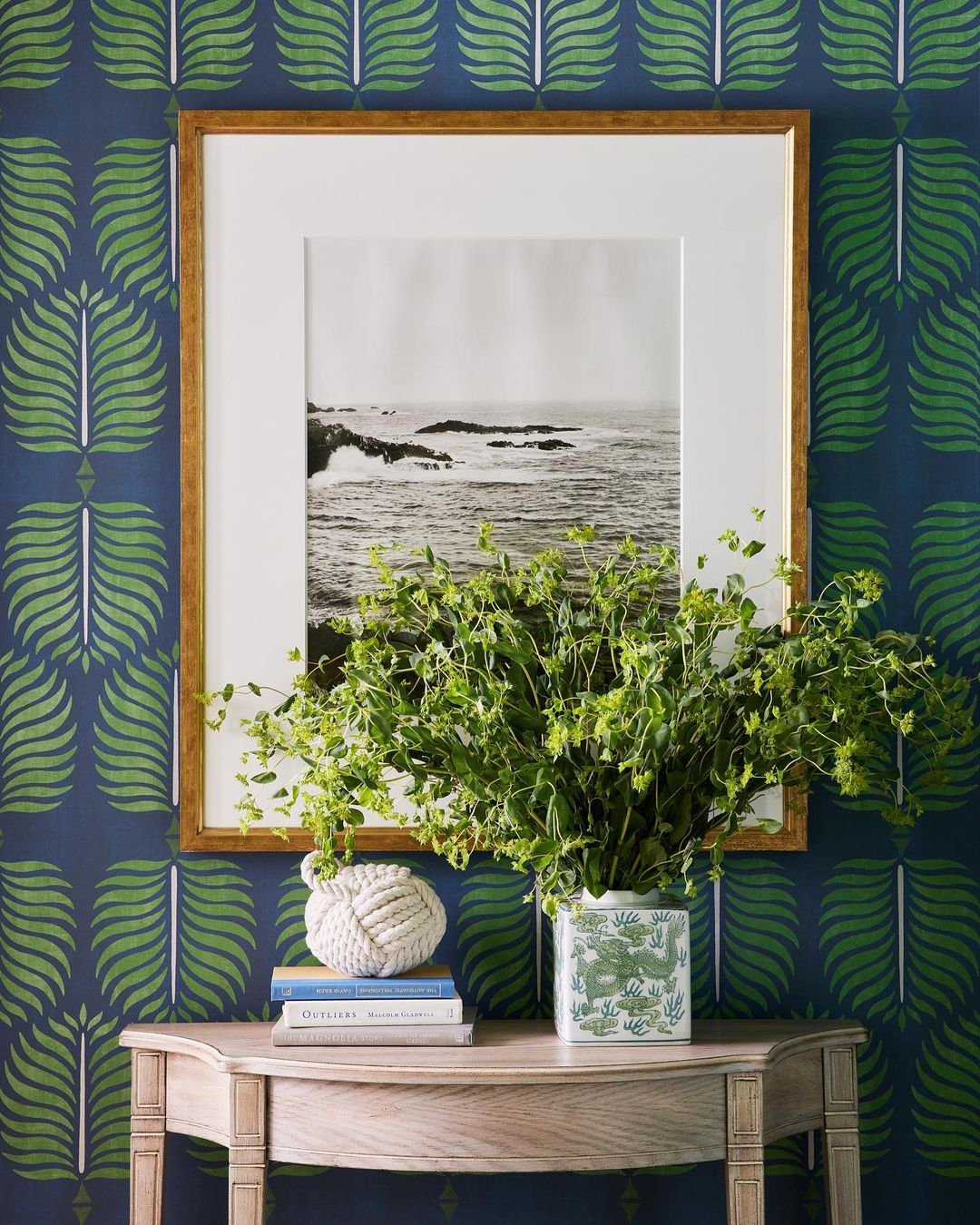 Interior decor with a wooden side table, vase of greenery, books, and framed ocean picture against patterned wallpaper.