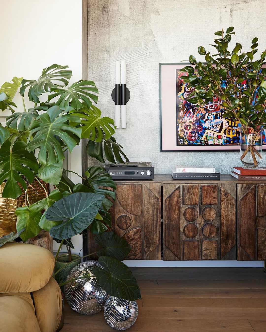 Eclectic interior featuring a wooden cabinet, plants, disco balls, and artwork against a textured wall, creating a visually rich living space.