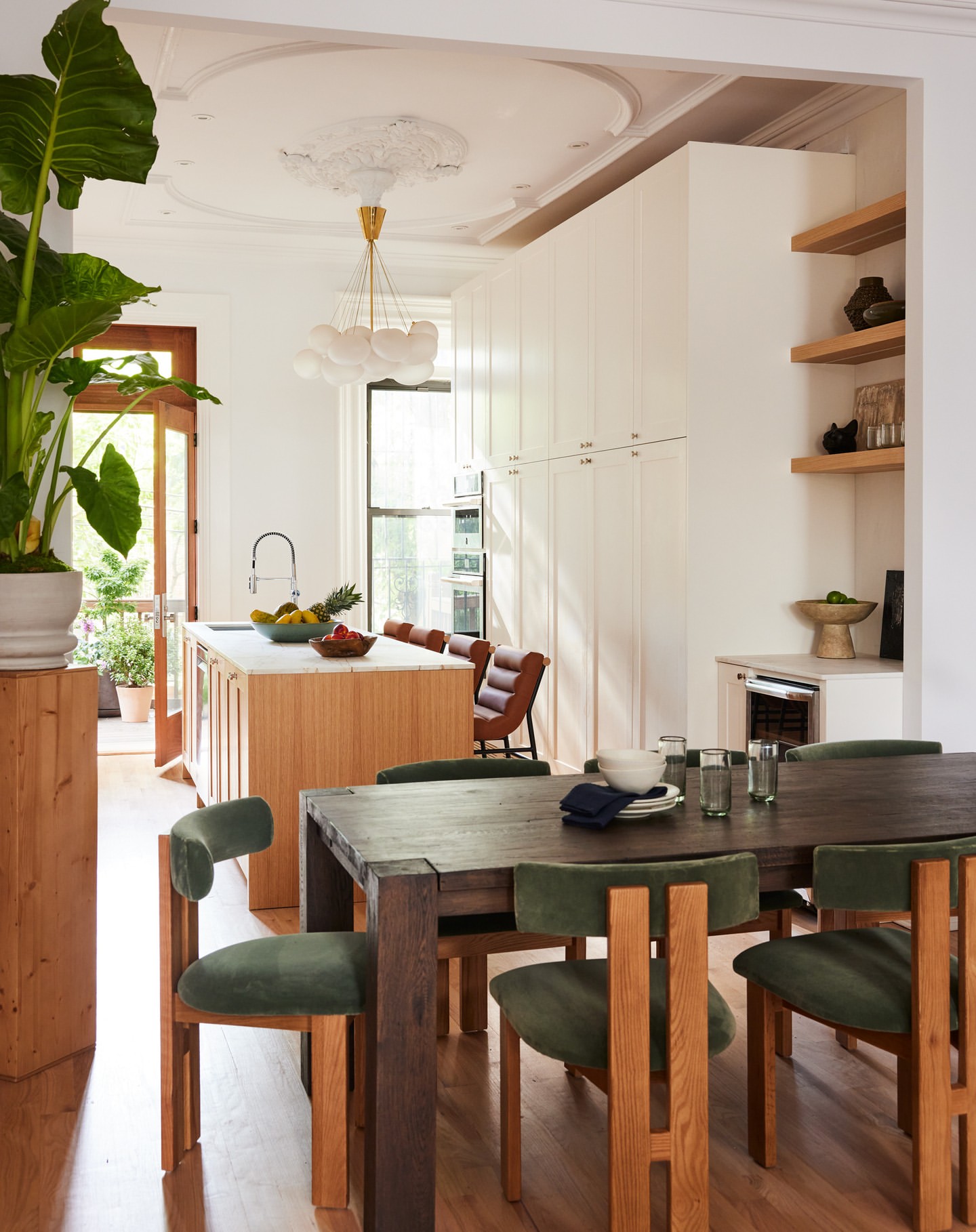 Interior design of a bright dining area with a large dark wood table, olive-green velvet chairs, and an open kitchen in the background.