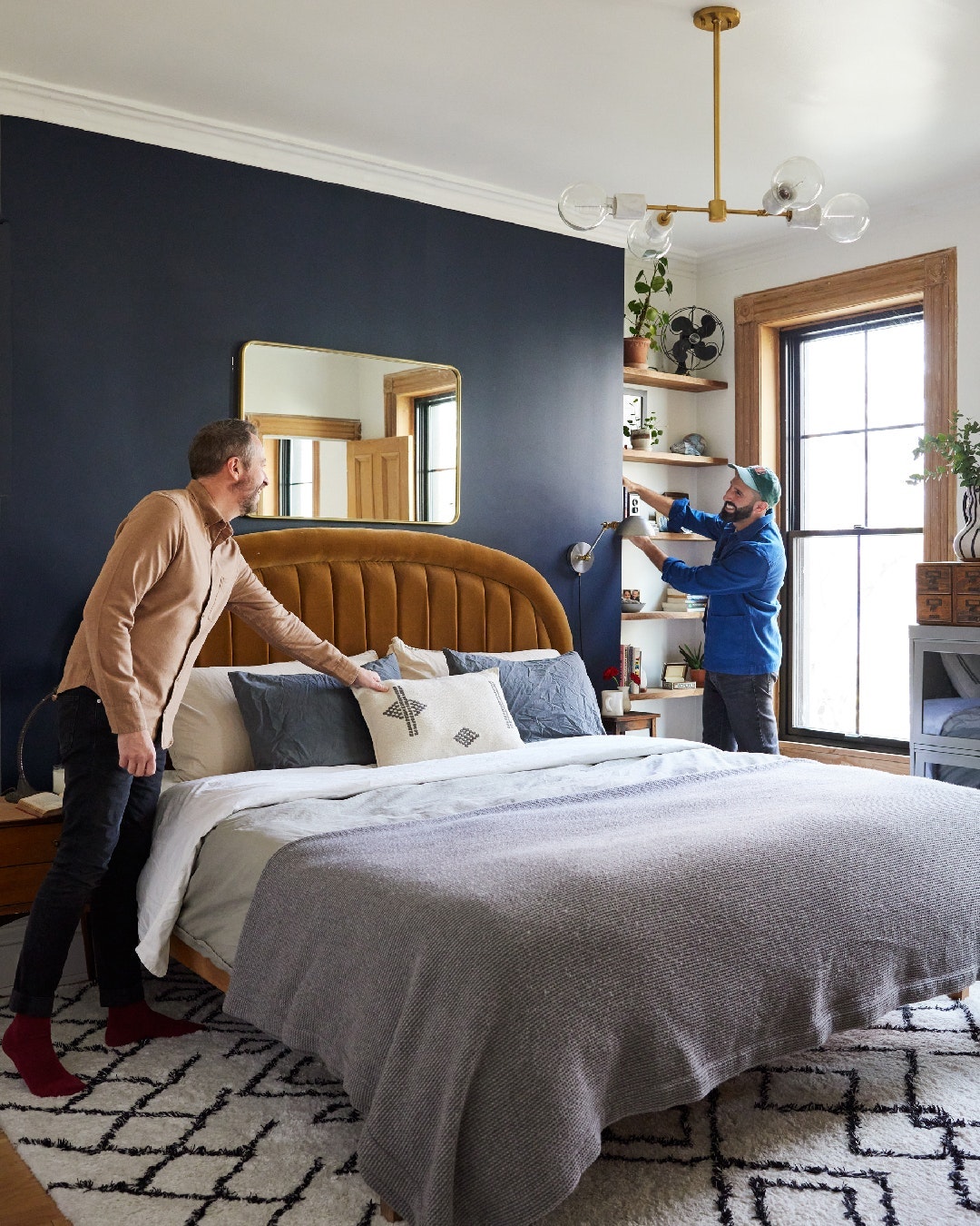 Two adult males in a bedroom with dark blue accent wall, gold light fixture, bed with headboard, neutral bedding, geometric patterned rug.