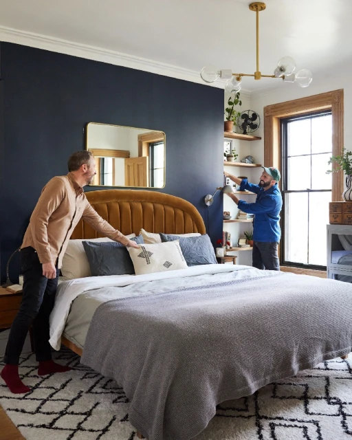 Two adult males in a bedroom with dark blue accent wall, gold light fixture, bed with headboard, neutral bedding, geometric patterned rug.