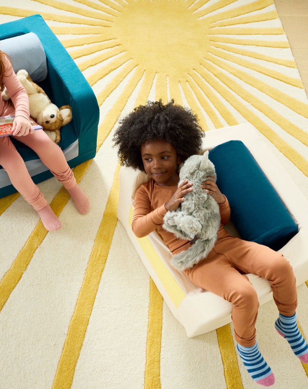 Child with afro lays on modular couch with a plush toy. Another child and teddy bear sit on matching couch near a sun-patterned rug.