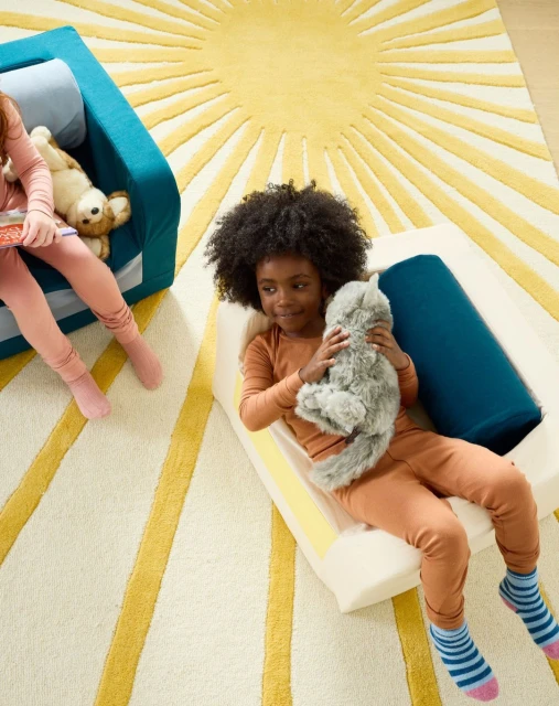 Child with afro lays on modular couch with a plush toy. Another child and teddy bear sit on matching couch near a sun-patterned rug.