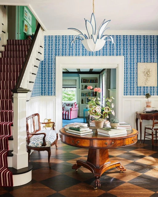 Elegant home foyer with patterned blue wallpaper, a round wood table topped with books and flowers, and a staircase with striped runner.