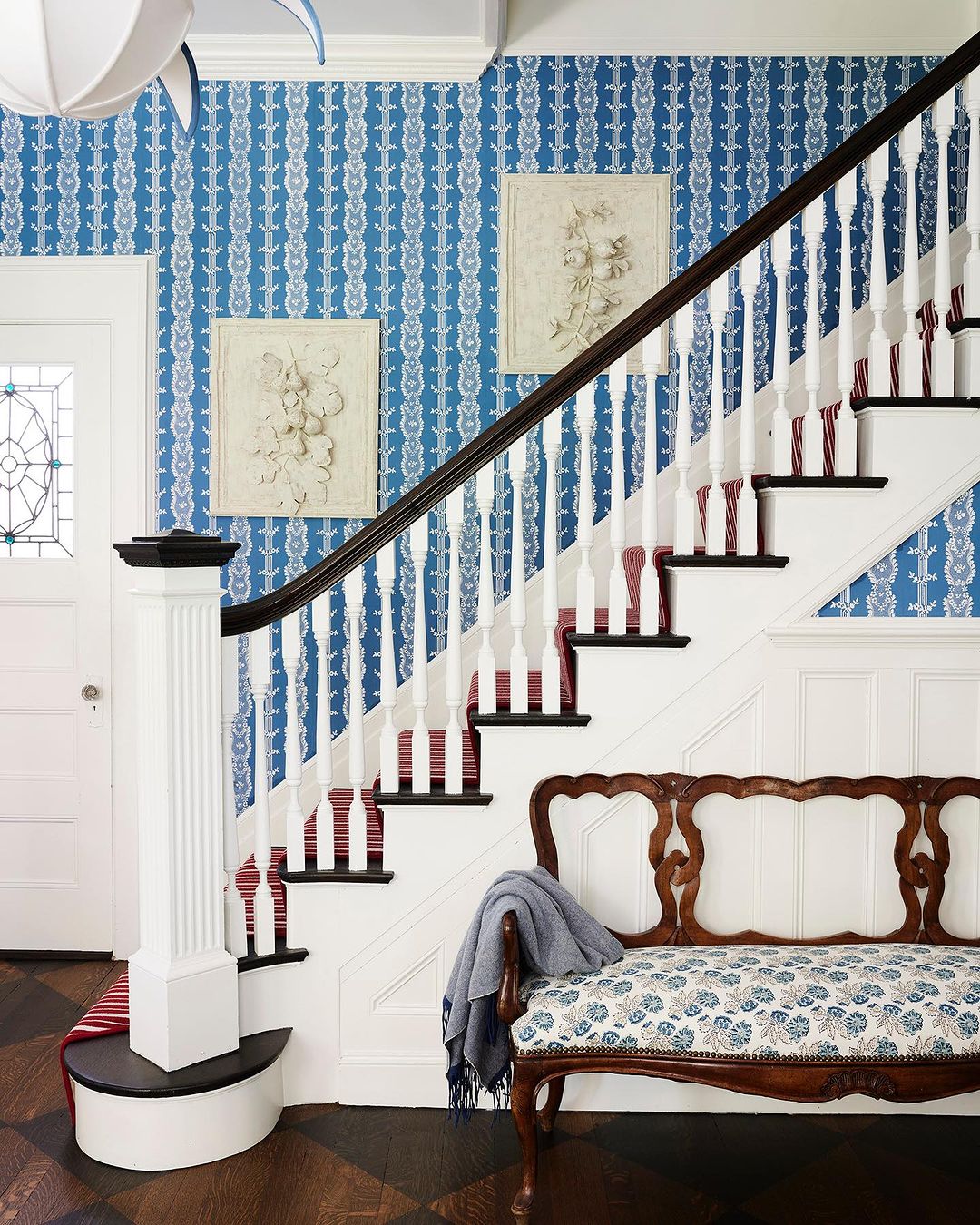 Staircase with floral wallpaper and framed art. The stairs are white with a dark brown banister and a patterned red carpet runner.