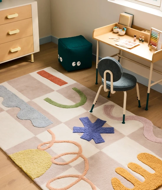 Interior of a child's room with a colorful rug, a wooden desk with objects, a chair, a green pouf, and a wooden dresser.