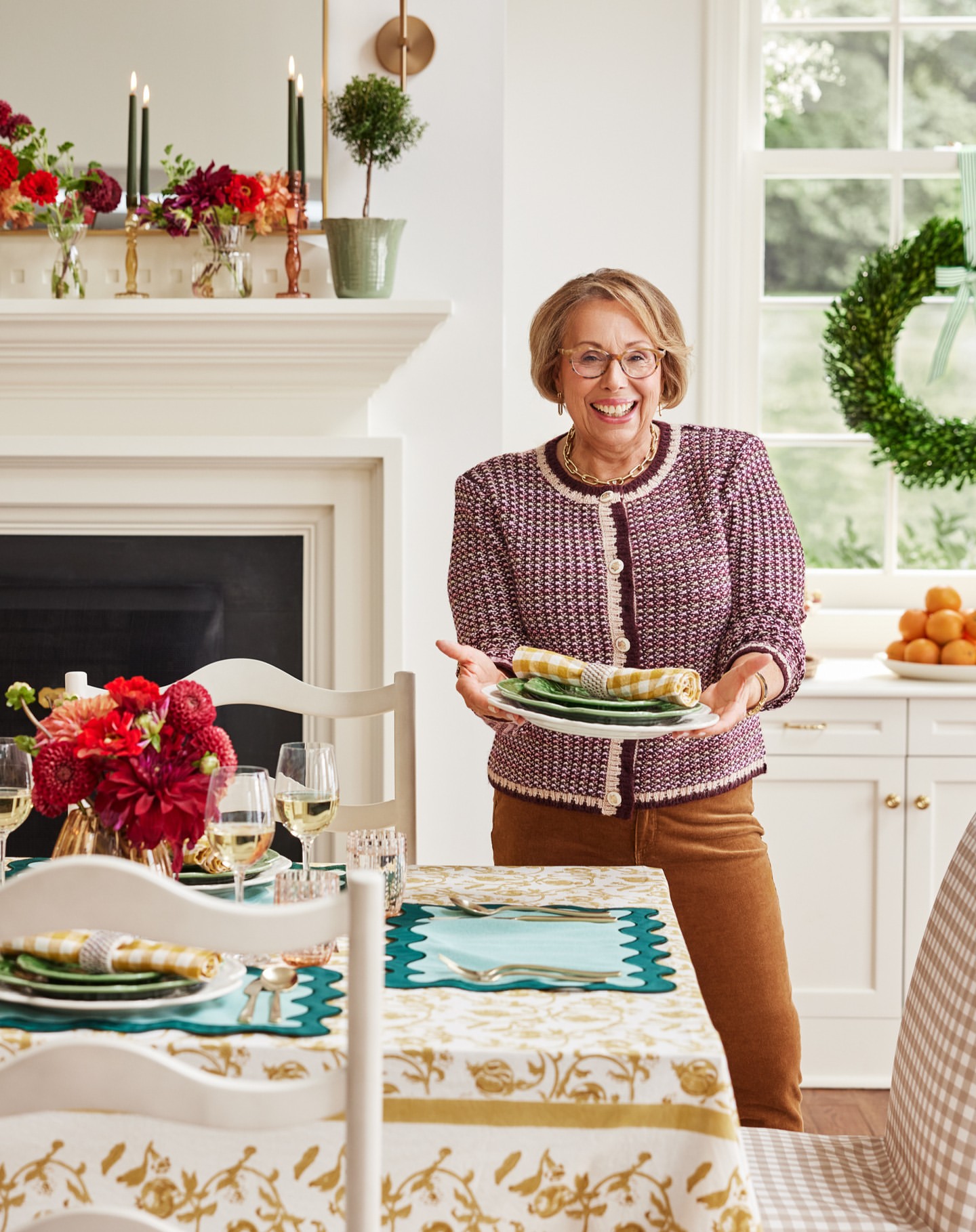 Smiling mature woman holding plates in a bright, stylish dining room with a floral tablecloth, dahlias, white wine glasses, and a wreath.